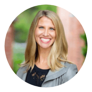 Professional smiling blonde woman in a gray blazer, set against a blurred green outdoor backdrop, framed in a circular border.