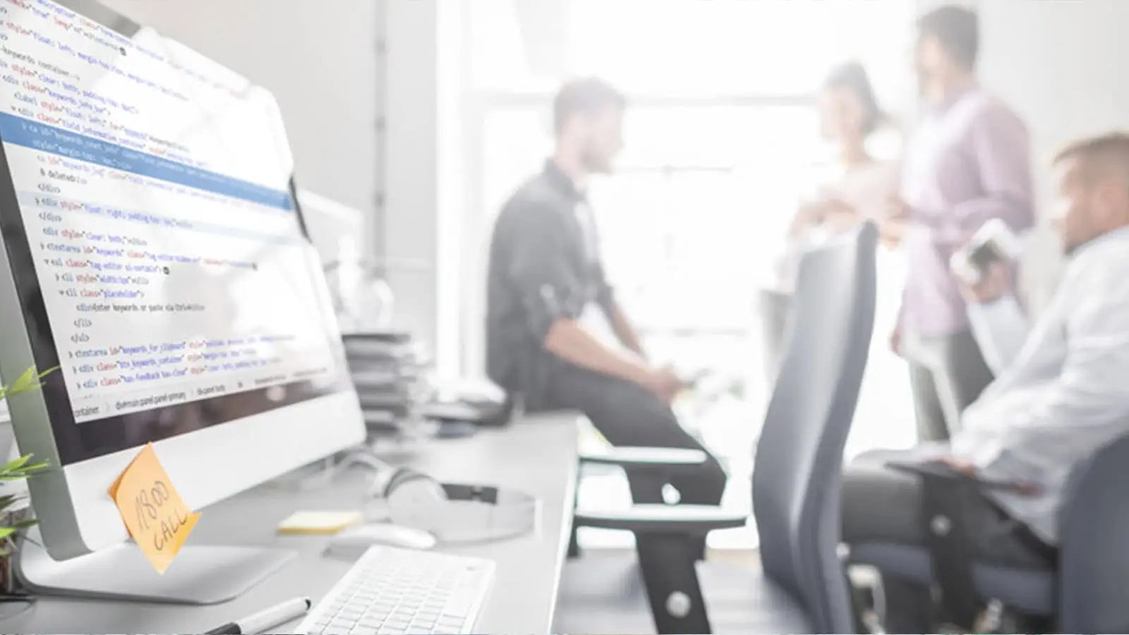 Modern office environment with a focus on a computer screen displaying code, surrounded by blurred professionals collaborating.