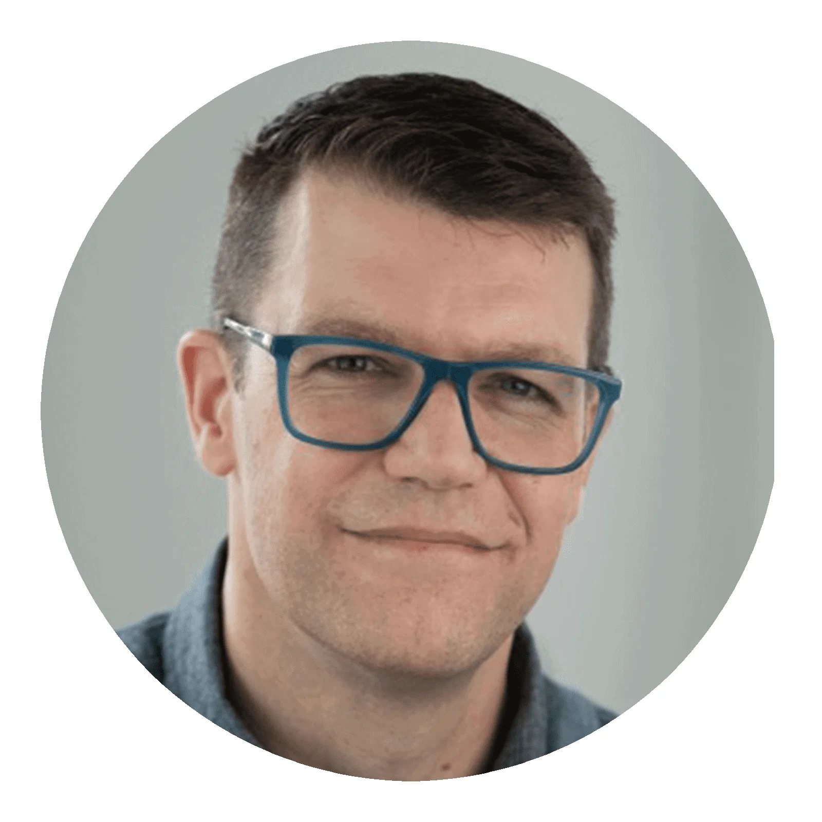 Professional man in blue glasses with confident smile, dressed in casual blue shirt, against a light gray background.