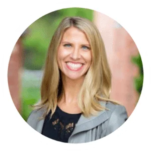 Professional smiling blonde woman in a gray blazer, set against a blurred green outdoor backdrop, framed in a circular border.