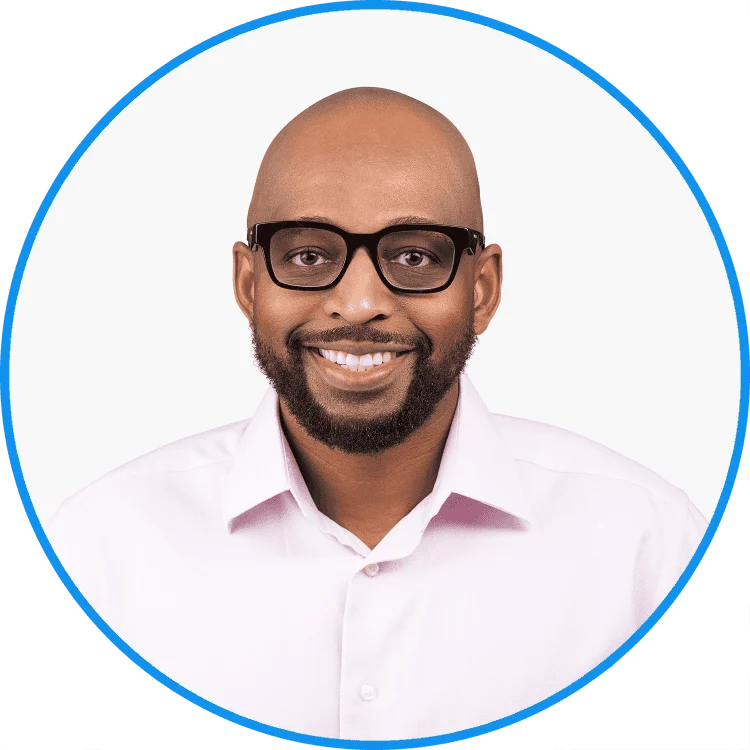 Professional African American businessman with glasses smiling, wearing a pink shirt against a white background.